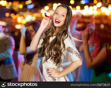 people, holidays and celebration concept - happy young woman or teen girl in fancy dress with sequins and party horn at night club over crowd and lights background
