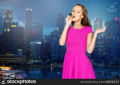 people, holidays and celebration concept - happy young woman or teen girl in pink dress blowing to party horn over night singapore city background