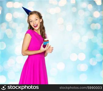 people, holidays and celebration concept - happy young woman or teen girl in pink dress and party cap with birthday cupcake over blue lights background