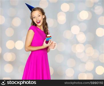 people, holidays and celebration concept - happy young woman or teen girl in pink dress and party cap with birthday cupcake over lights background