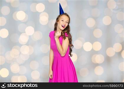 people, holidays and celebration concept - happy young woman or teen girl in pink dress and party cap over lights background