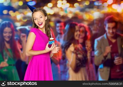 people, holidays and celebration concept - happy young woman or teen girl in pink dress and party cap with birthday cupcake at disco club over crowd lights background