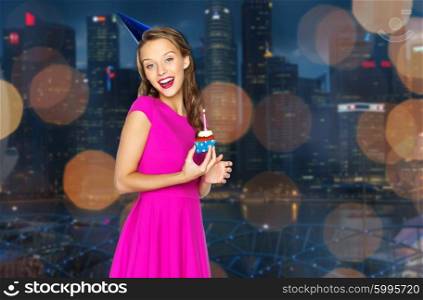 people, holidays and celebration concept - happy young woman or teen girl in pink dress and party cap with birthday cupcake over night singapore city lights background