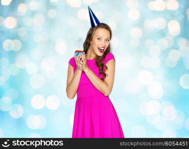 people, holidays and celebration concept - happy young woman or teen girl in pink dress and party cap holding birthday cupcake with burning candle over blue holidays lights background