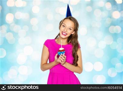 people, holidays and celebration concept - happy young woman or teen girl in pink dress and party cap holding birthday cupcake with burning candle over blue holidays lights background