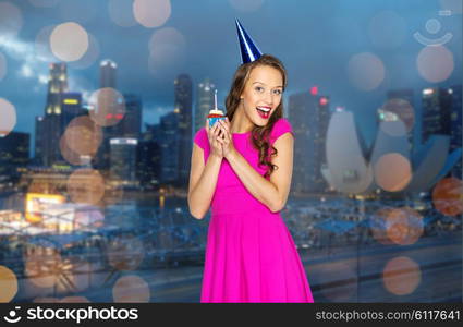 people, holidays and celebration concept - happy young woman or teen girl in pink dress and party cap holding birthday cupcake with burning candle