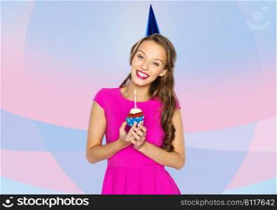 people, holidays and celebration concept - happy young woman or teen girl in pink dress and party cap holding birthday cupcake with burning candle over pink violet background