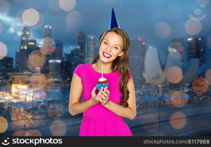 people, holidays and celebration concept - happy young woman or teen girl in pink dress and party cap holding birthday cupcake with burning candle