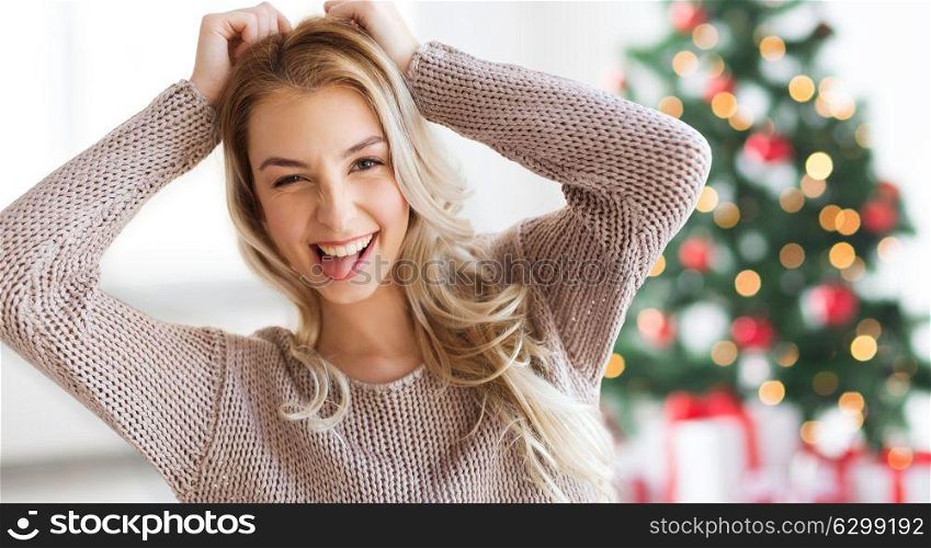 people, holidays and emotion concept - happy young woman or teenage girl having fun and showing tongue at home over christmas tree background. happy young woman or teenage girl at christmas