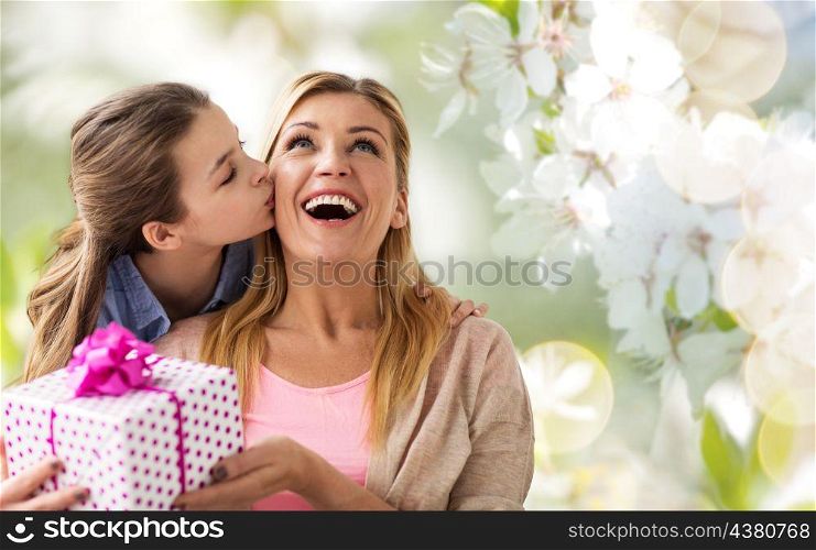 people, holidays and family concept - daughter kissing happy mother and giving her birthday present over cherry blossom background. daughter kissing mother and giving her present. daughter kissing mother and giving her present