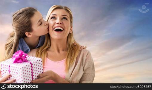 people, holidays and family concept - daughter kissing happy mother and giving her birthday present over evening sky background. daughter kissing mother and giving her present. daughter kissing mother and giving her present