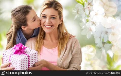 people, holidays and family concept - daughter kissing happy mother and giving her birthday present over cherry blossom background. daughter kissing mother and giving her present
