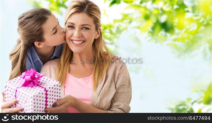 people, holidays and family concept - daughter kissing happy mother and giving her birthday present over green natural background. daughter kissing mother and giving her present. daughter kissing mother and giving her present