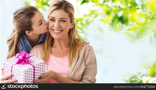people, holidays and family concept - daughter kissing happy mother and giving her birthday present over green natural background. daughter kissing mother and giving her present. daughter kissing mother and giving her present