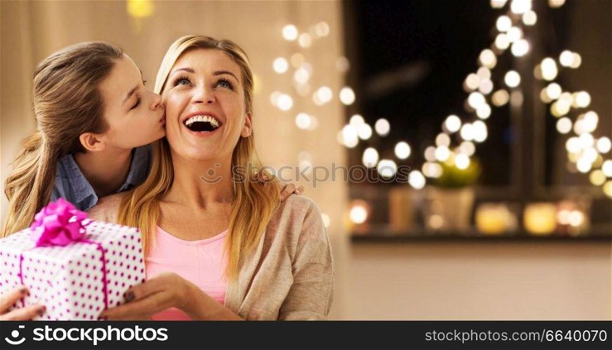 people, holidays and family concept - happy daughter giving birthday present and kissing her mother at home over garland lights background. daughter giving present to mother on christmas