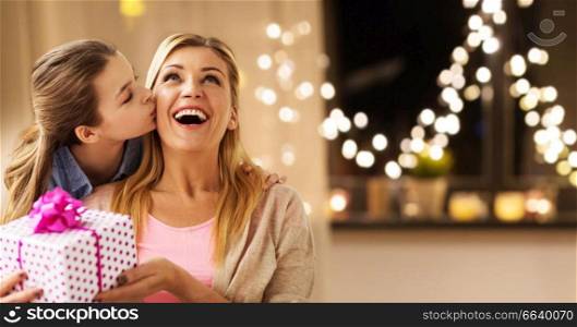 people, holidays and family concept - happy daughter giving birthday present and kissing her mother at home over garland lights background. daughter giving present to mother on christmas