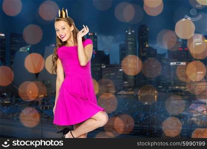 people, holidays and fashion concept - happy young woman or teen girl in pink dress and princess crown over city and lights background