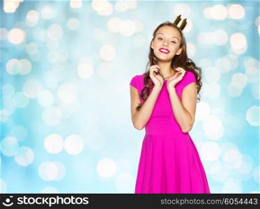 people, holidays and fashion concept - happy young woman or teen girl in pink dress and princess crown over blue holidays lights background