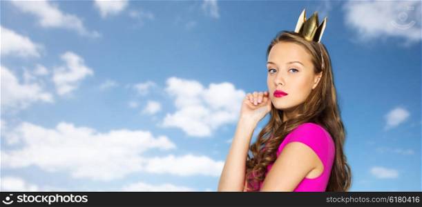 people, holidays and fashion concept - young woman or teen girl in pink dress and princess crown over blue sky and clouds background