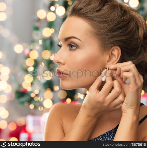 people, holidays and glamour concept - close up of beautiful woman wearing earrings over christmas tree and lights background