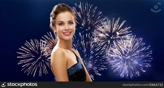people, holidays and glamour concept - happy beautiful woman in evening dress over firework on dark blue background