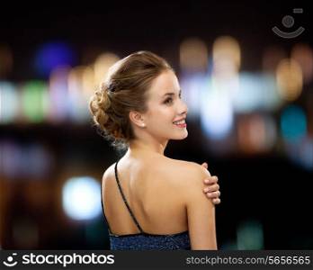people, holidays and glamour concept - smiling woman in evening dress over black background over night lights background from back