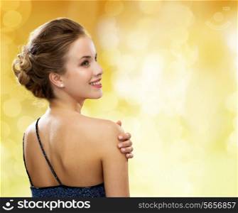 people, holidays and glamour concept - smiling woman in evening dress over black background over yellow lights background from back