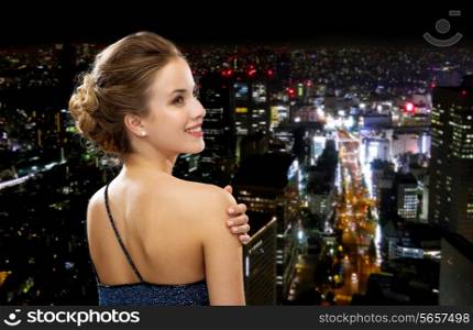 people, holidays and glamour concept - smiling woman in evening dress over black background over night city background from back