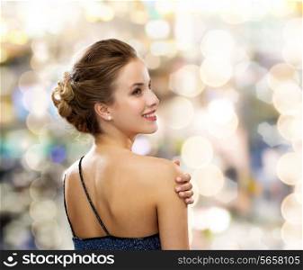 people, holidays and glamour concept - smiling woman in evening dress over black background over lights background from back