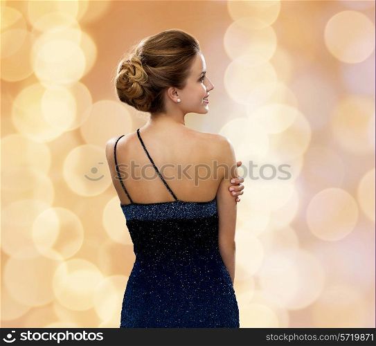 people, holidays and glamour concept - smiling woman in evening dress over beige lights background from back