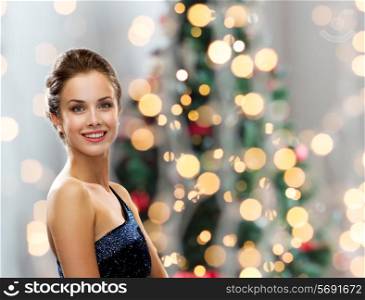 people, holidays and glamour concept - smiling woman in evening dress over christmas tree lights background