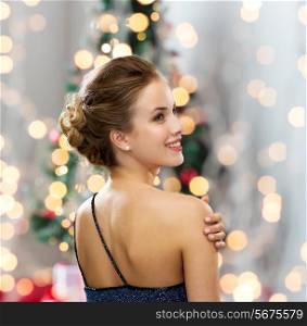 people, holidays, and glamour concept - smiling woman in evening dress showing earrings over christmas tree and lights background