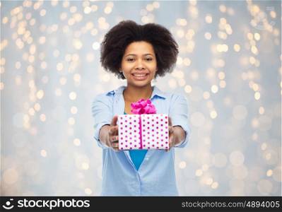 people, holidays and greeting concept - happy african american young woman with birthday gift box over holidays lights background