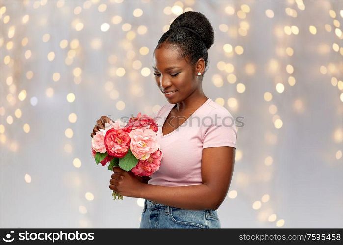 people, holidays and international women&rsquo;s day concept - happy african american young woman with bunch of peony flowers over festive lights background. happy african american woman with bunch of flowers