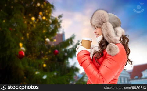 people, holidays and leisure concept - happy woman in winter fur with coffee cup hat over christmas tree at tallinn old town hall square background. woman with coffee over christmas tree in tallinn