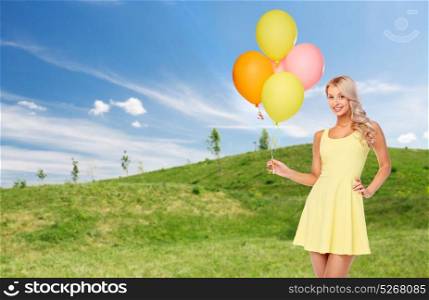 people, holidays and summer party concept - happy young woman or teen girl in yellow dress with helium air balloons over blue sky and green field background. happy woman in summer dress with air balloons