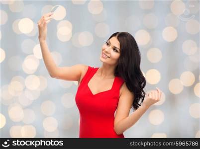people, holidays and technology concept - beautiful sexy woman in red dress taking selfie picture by smartphone over lights background
