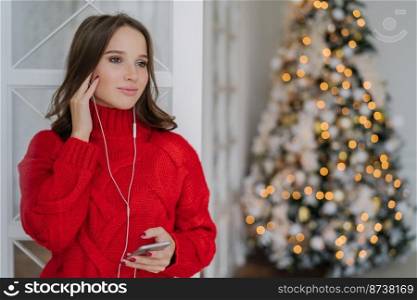 People, holidays and technology concept. Pretty woman uses mobile phone and earphones for listening music, stands at home against Christmas tree lights background with copy space for your text