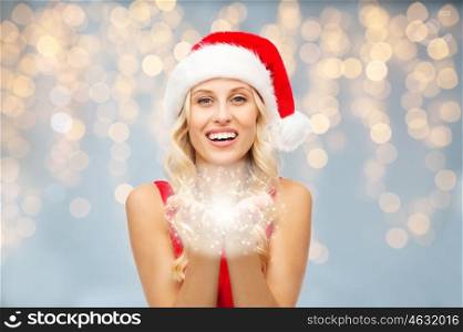 people, holidays, christmas and advertisement concept - happy blonde woman in santa hat holding fairy dust in her hands over lights background