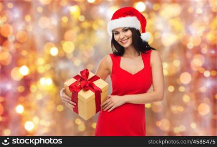 people, holidays, christmas and celebration concept - beautiful sexy woman in red dress and santa hat with gift box over lights background