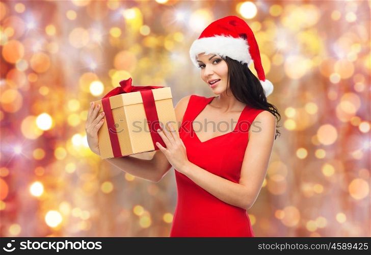 people, holidays, christmas and celebration concept - beautiful sexy woman in red dress and santa hat with gift box over lights background
