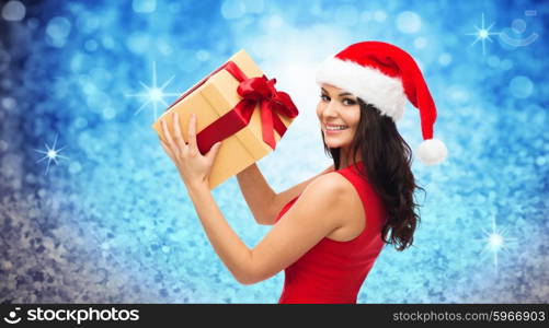people, holidays, christmas and celebration concept - beautiful sexy woman in red dress and santa hat with gift box over blue glitter or lights background