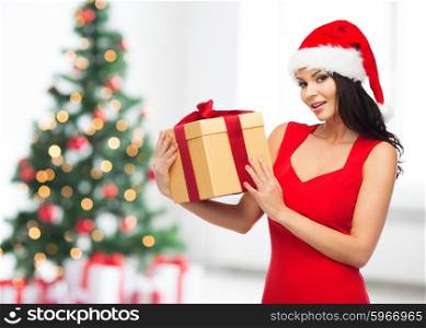 people, holidays, christmas and celebration concept - beautiful sexy woman in red dress and santa hat with gift box over room with christmas tree and gifts background