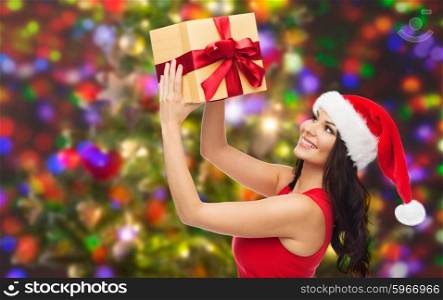 people, holidays, christmas and celebration concept - beautiful sexy woman in red dress and santa hat with gift box over party lights background