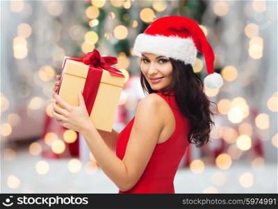 people, holidays, christmas and celebration concept - beautiful sexy woman in red dress and santa hat with gift box over christmas tree lights and presents background