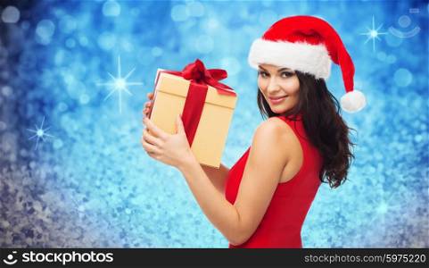 people, holidays, christmas and celebration concept - beautiful sexy woman in red dress and santa hat with gift box over blue glitter and lights background