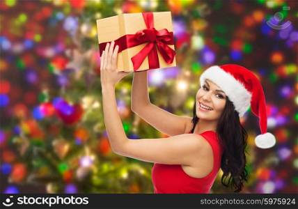 people, holidays, christmas and celebration concept - beautiful sexy woman in red dress and santa hat with gift box over holidays lights background