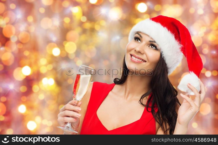 people, holidays, christmas and celebration concept - beautiful sexy woman in santa hat and red dress with champagne glass over lights background