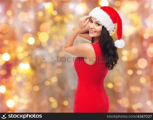people, holidays, christmas and celebration concept - beautiful sexy woman in santa hat and red dress over lights background