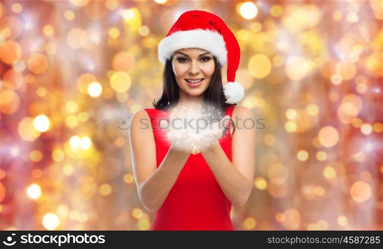 people, holidays, christmas and celebration concept - beautiful sexy woman in santa hat and red dress holding fairy dust or snowflakes on her palms over lights background